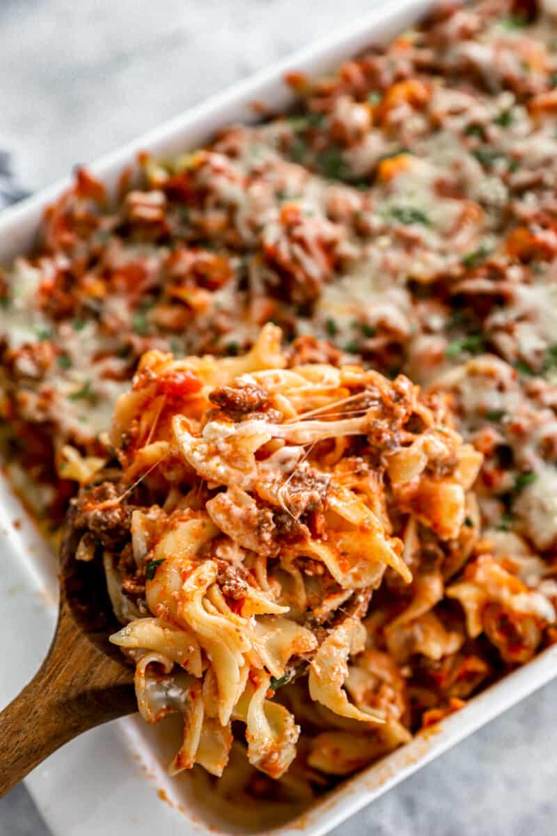 close up of a wooden spoon lifting a scoop of sour cream noodle casserole from a rectangular baking dish.