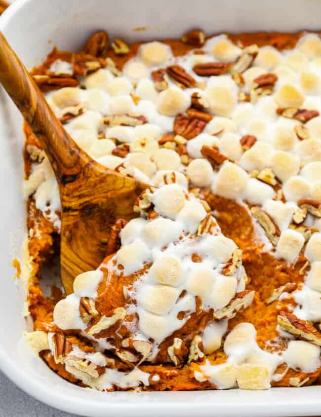 a wooden spoon scooping sweet potato casserole from a white rectangular baking dish.