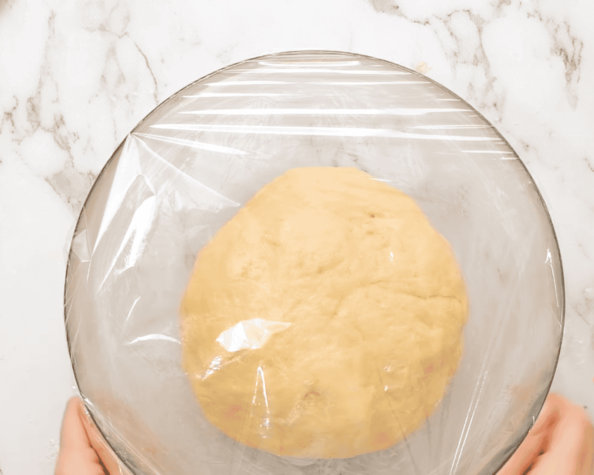 Letting the dough rise in a bowl covered with plastic wrap.