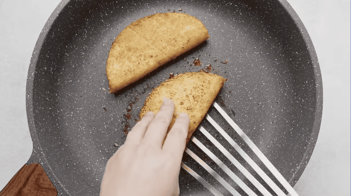 Two golden-brown, crispy birria tacos being cooked in a speckled nonstick frying pan. A hand is gently pressing down on one taco with a metal spatula, ensuring even crispiness.