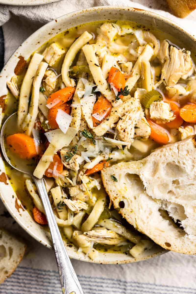 overhead view of a serving of homemade chicken noodle soup in a white bowl with a spoon and a slice of bread.