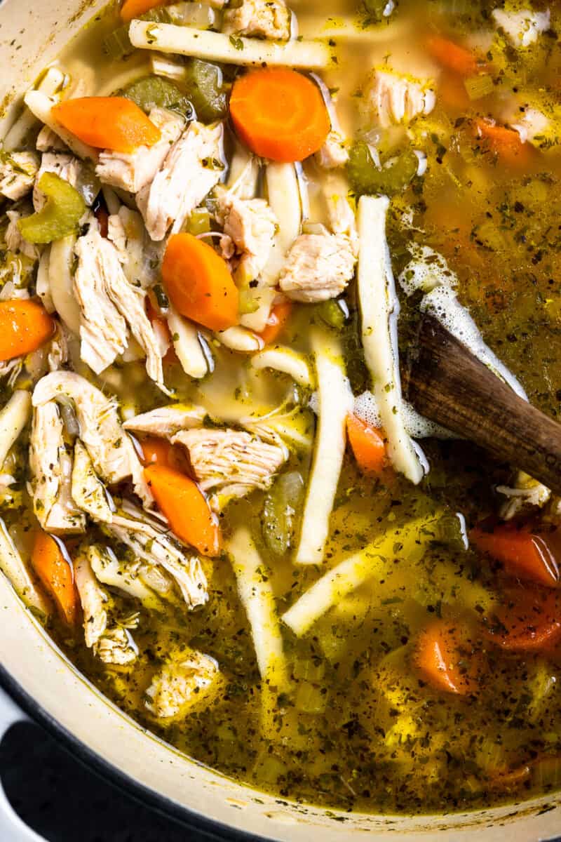 overhead close up view of homemade chicken noodle soup in a white dutch oven with a wooden spoon.