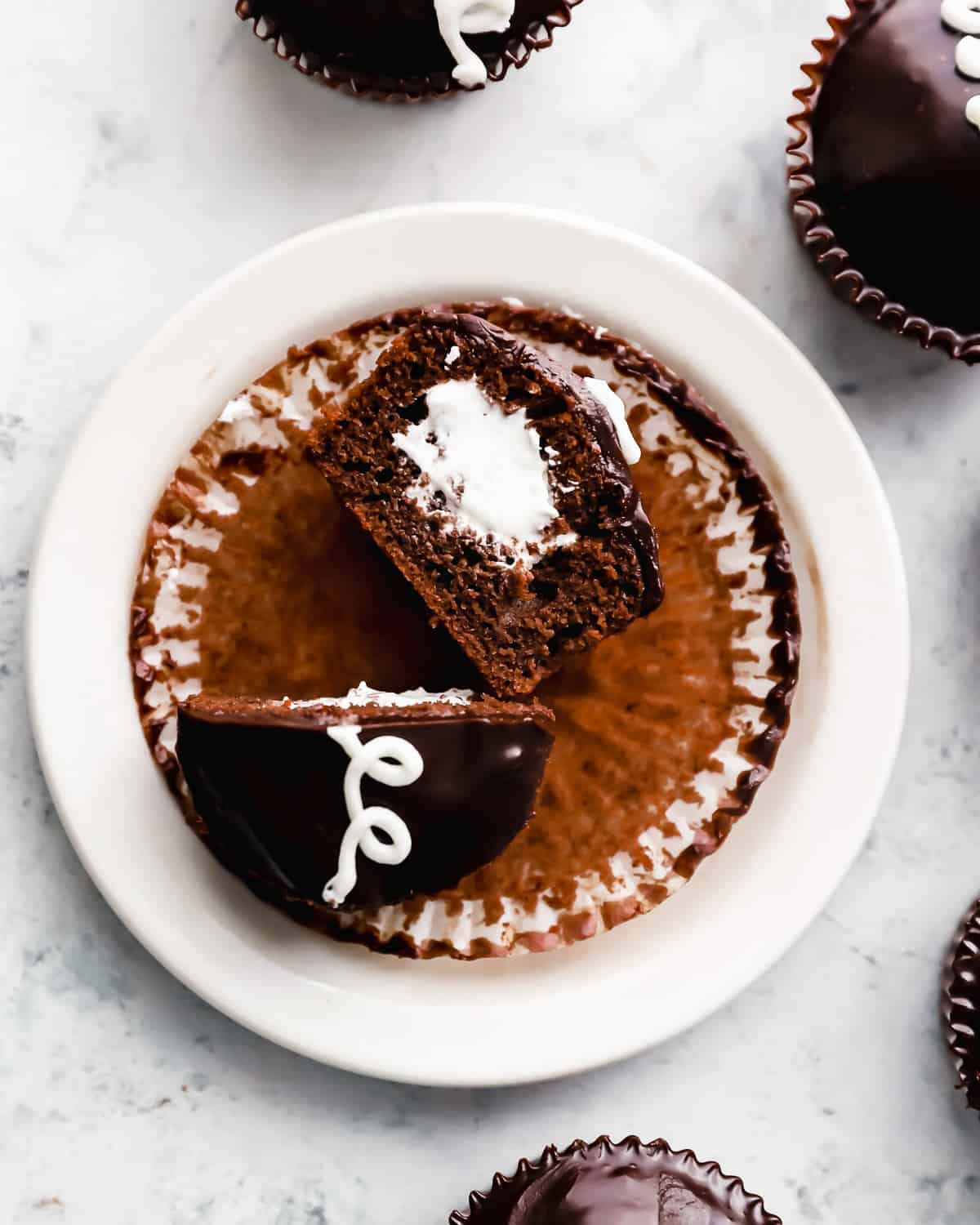 overhead view of a halved copycat hostess cupcake on a cupcake liner on a white plate, with one half upturned to show the fluffy creme filling, and the other upright to show the squiggle.