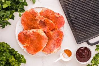 overhead view of 4 seasoned raw pork chops on a white plate.