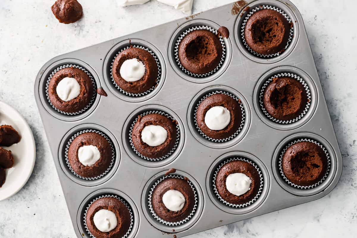 overhead view of 12 cored chocolate cupcakes in a cupcake tin, 8 of them have been filled with marshmallow creme.