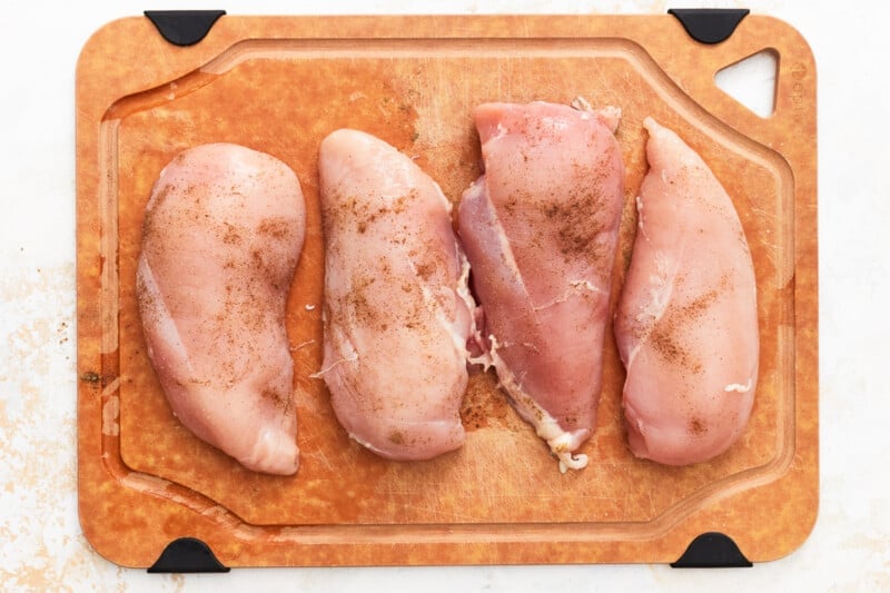 4 seasoned raw chicken breasts on a wooden cutting board.