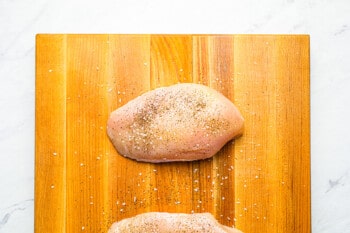 2 raw seasoned chicken breasts on a wooden cutting board.