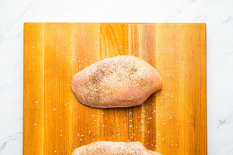 2 raw seasoned chicken breasts on a wooden cutting board.