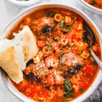 overhead view of a serving of meatball soup in a white bowl with a spoon and a piece of bread.