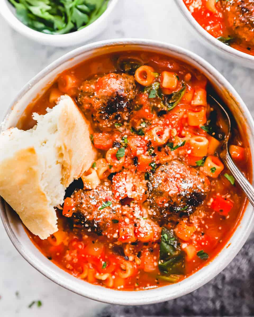 overhead view of a serving of meatball soup in a white bowl with a spoon and a piece of bread.