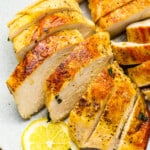 three-quarters view of 2 sliced sous vide chicken breasts on a white plate with a lemon slice.