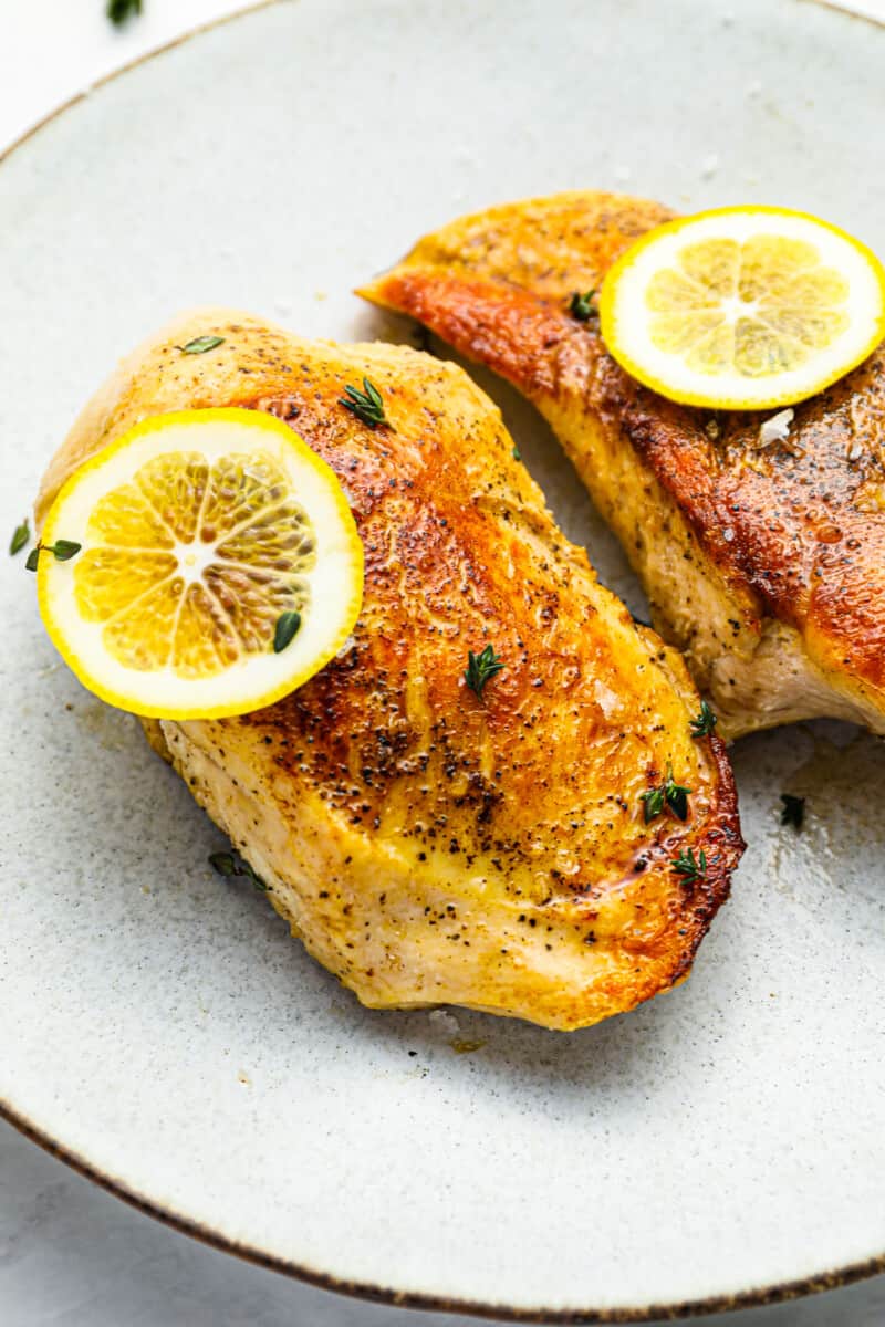 three-quarters view of 2 seared sous vide chicken breasts on a white plate topped with lemon slices.