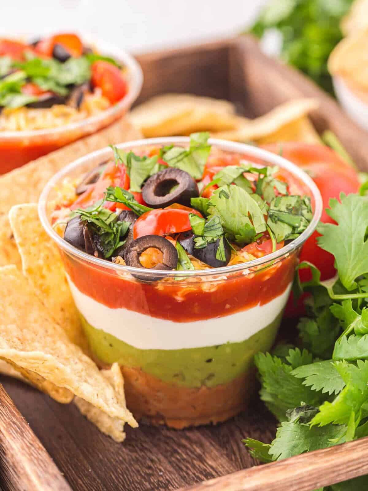 a 7 layer dip cup on a wooden tray with tortilla chips.