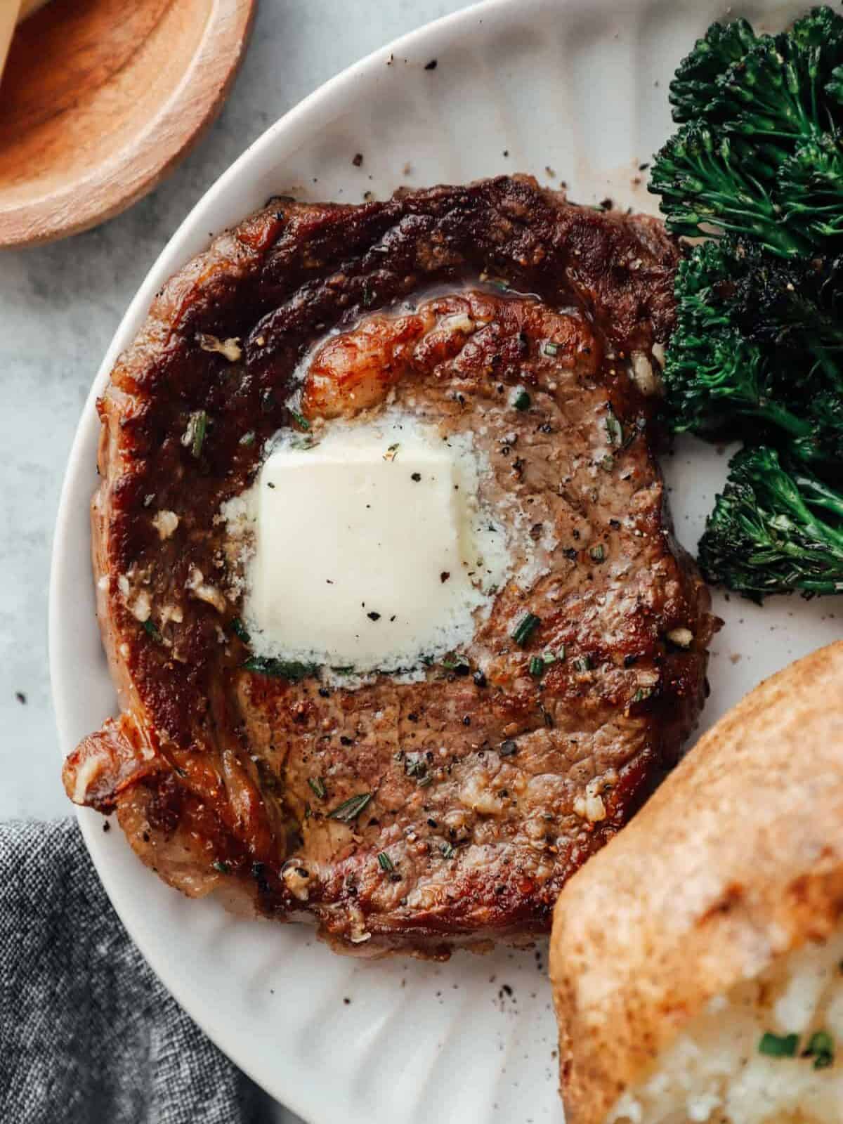 close up of a pan seared ribeye on a white plate with butter on top and a side of baked potato and broccoli rabe.