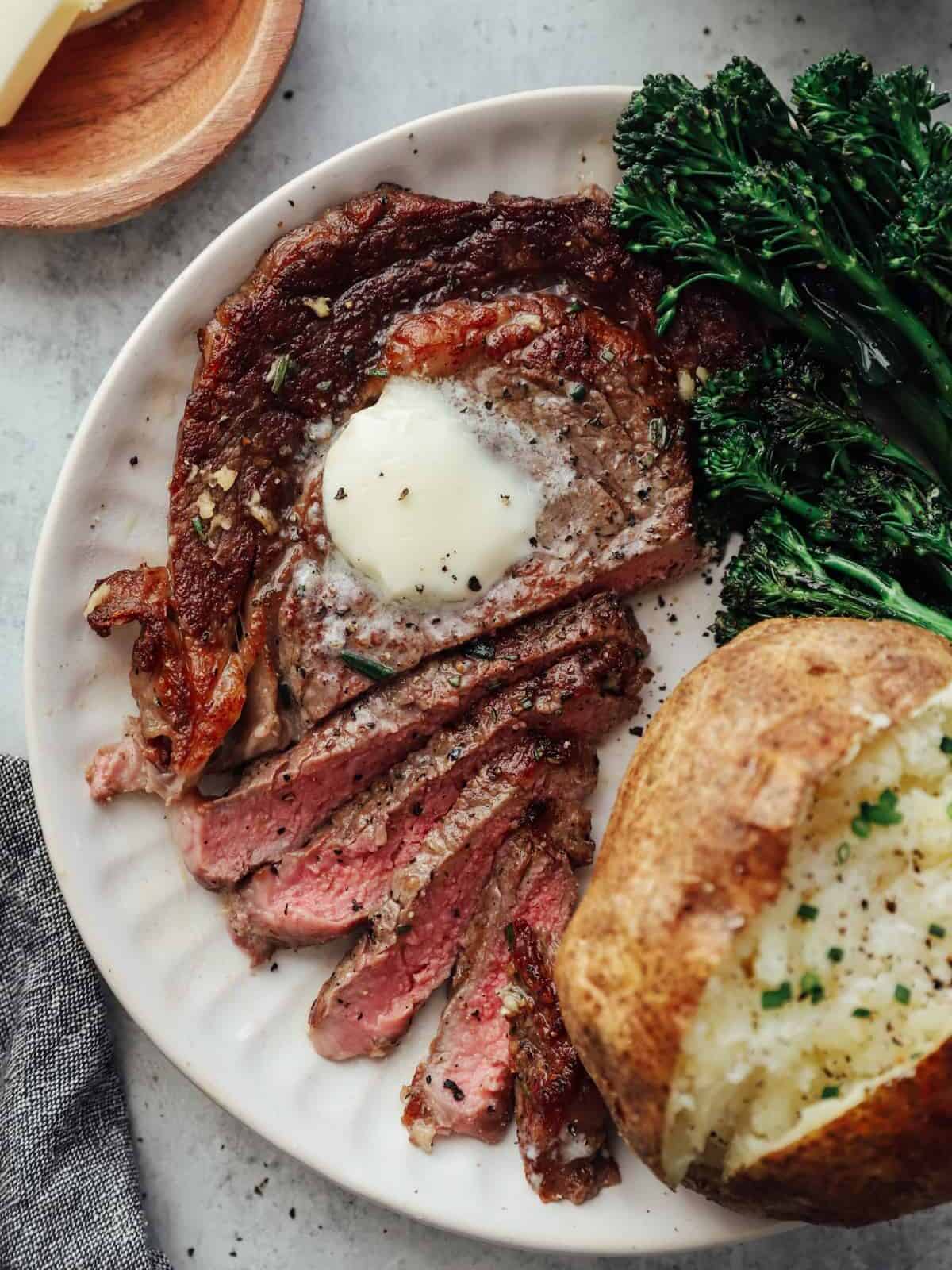 overhead view of a sliced pan seared ribeye on a white plate topped with butter and a side of baked potato and broccoli rabe.