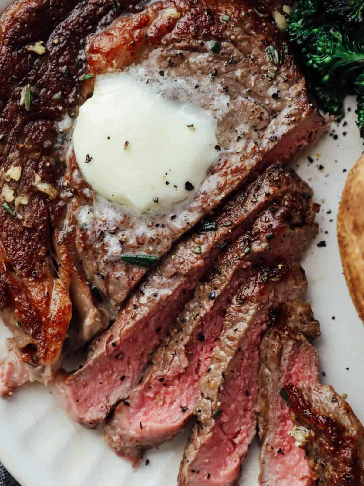 close up view of a sliced pan seared ribeye on a white plate with butter on top.