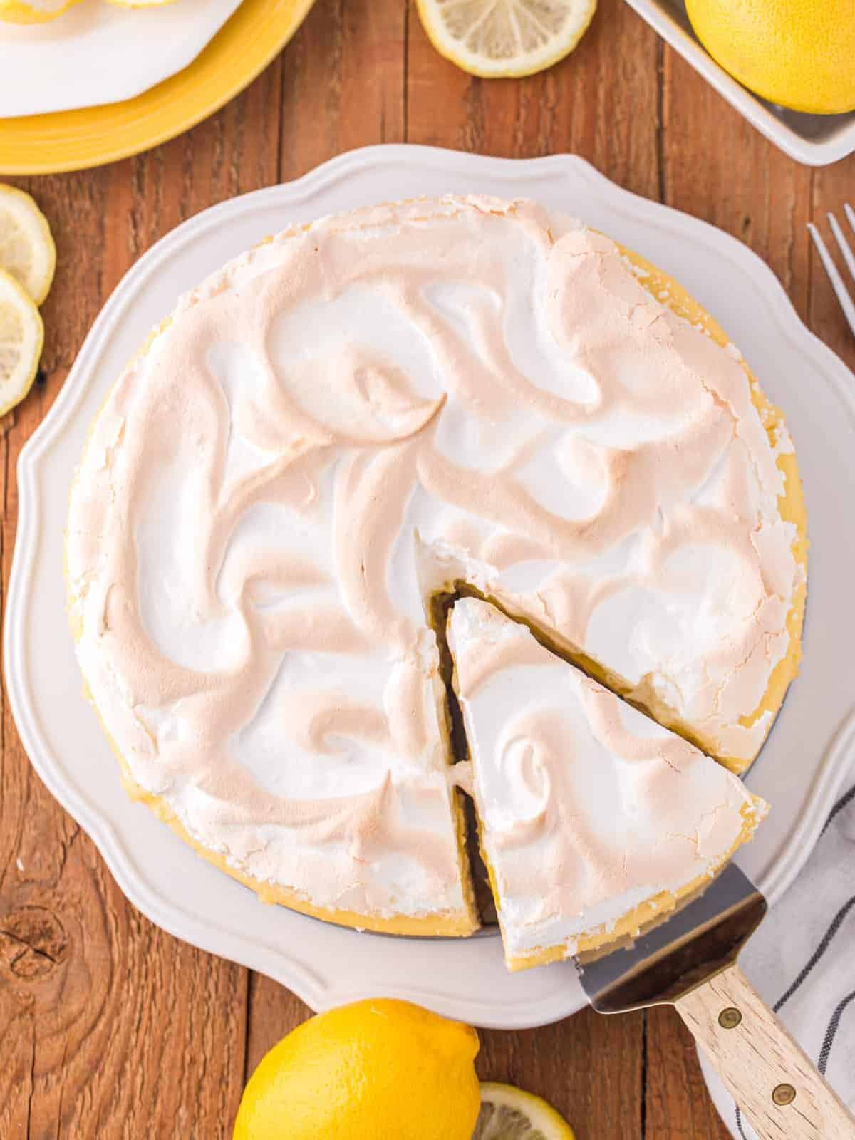 overhead view of a cake server removing a slice of lemon meringue cheesecake from a white cake stand.