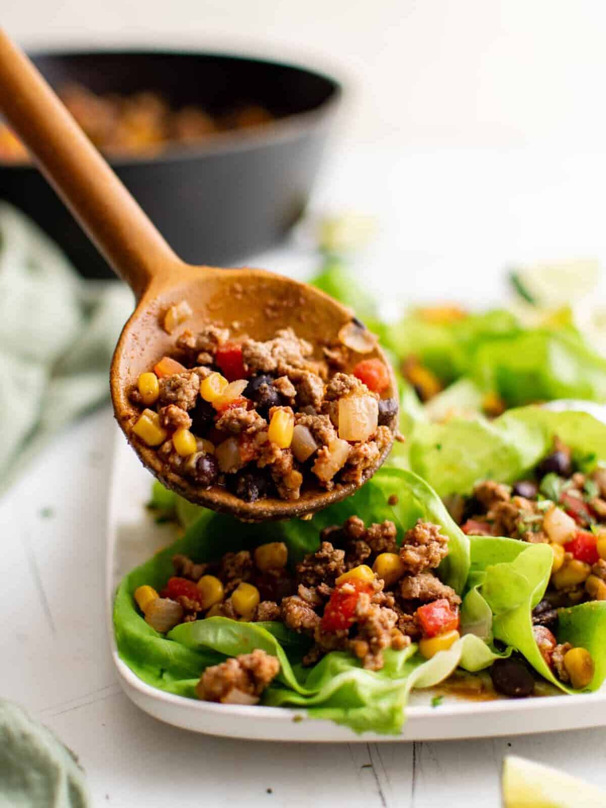 a spoonful of taco lettuce wrap meat (ground turkey with corn, onions, and black beans) held above a platter of lettuce wraps