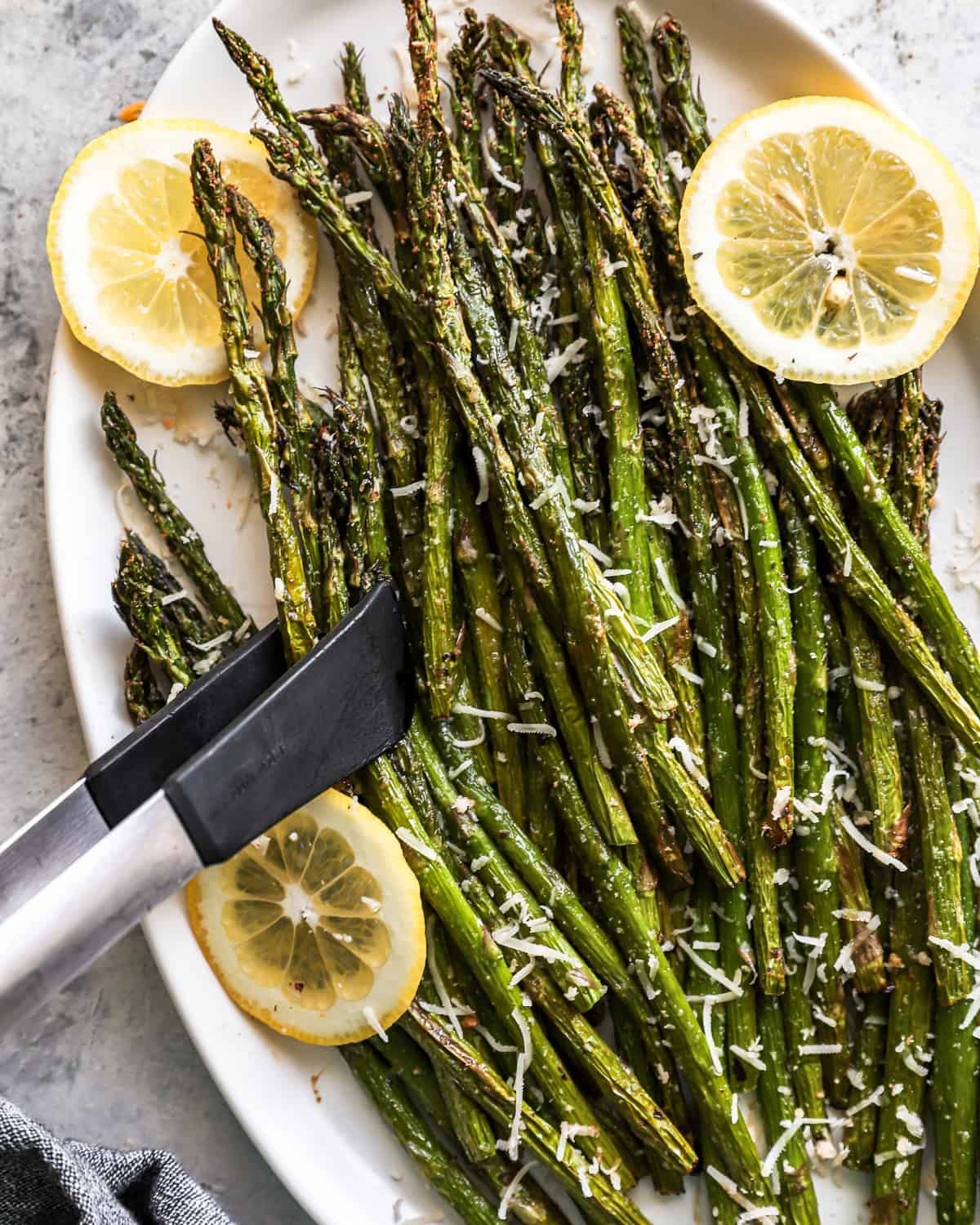 overhead view of tongs grabbing air fried asparagus from a white oval serving platter.