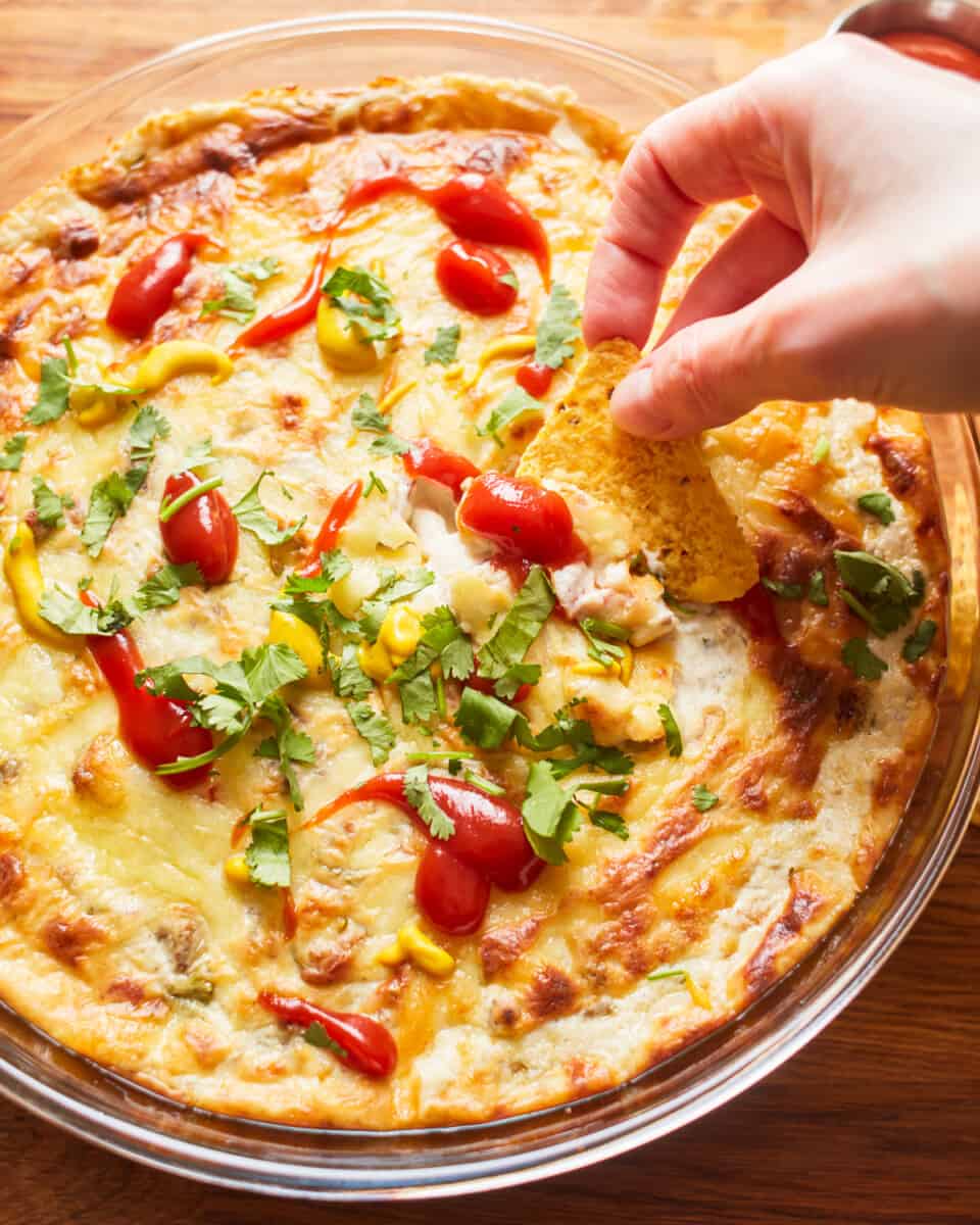 Cheeseburger Dip is the best dip recipe to celebrate Memorial Day and the Fourth of July! This bacon cheeseburger dip is loaded with bacon, Swiss, cheddar, hamburger, and everything else that makes cheeseburgers one of America's favorite foods! three-quarters view of a hand dipping a tortilla chip into cheeseburger dip in a round baking dish.