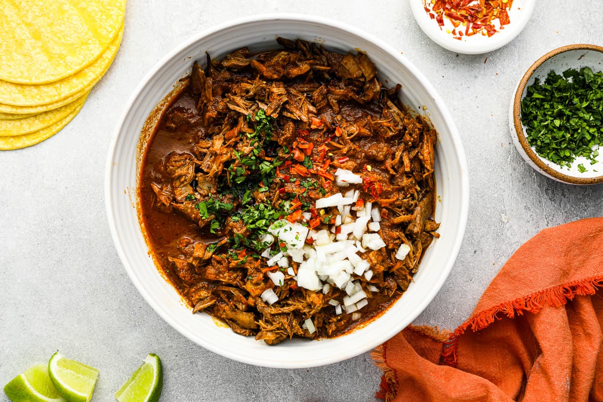 birria meat in a white bowl with limes and tortillas.