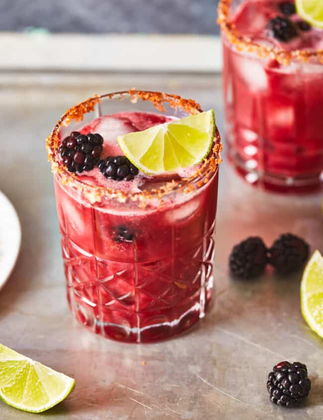 three-quarters view of a blackberry margarita in a short glass with a chili salt rim and blackberry and lime wedge garnish.