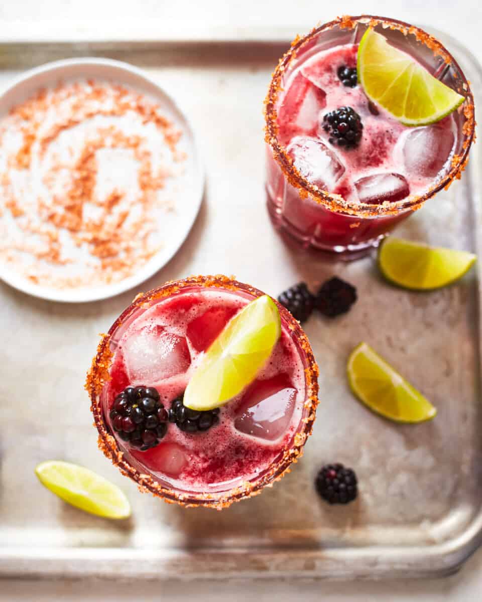 overhead view of 2 blackberry margaritas on a metal tray next to a small white plate filled with chili rimming salt.