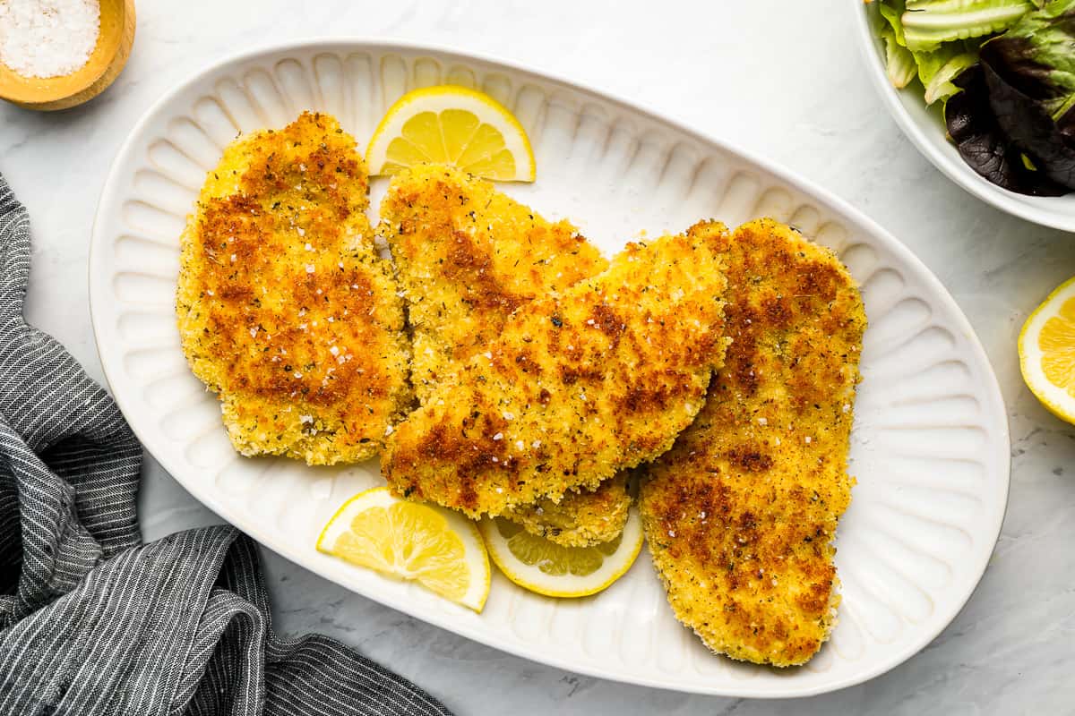 fried chicken breast cutlets on a white plate with lemon wedges.
