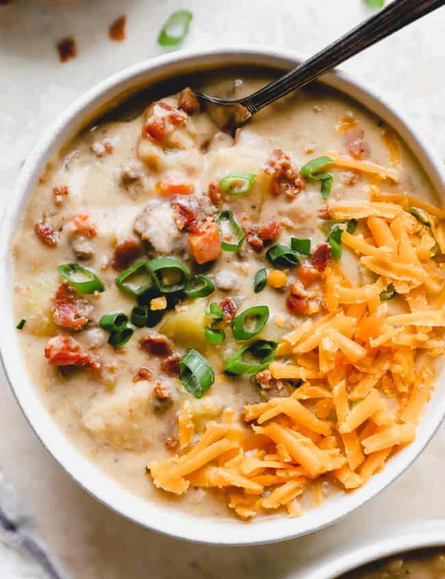 overhead view of a serving of cheeseburger soup in a white bowl with a spoon topped with bacon, green onion, and shredded cheese.