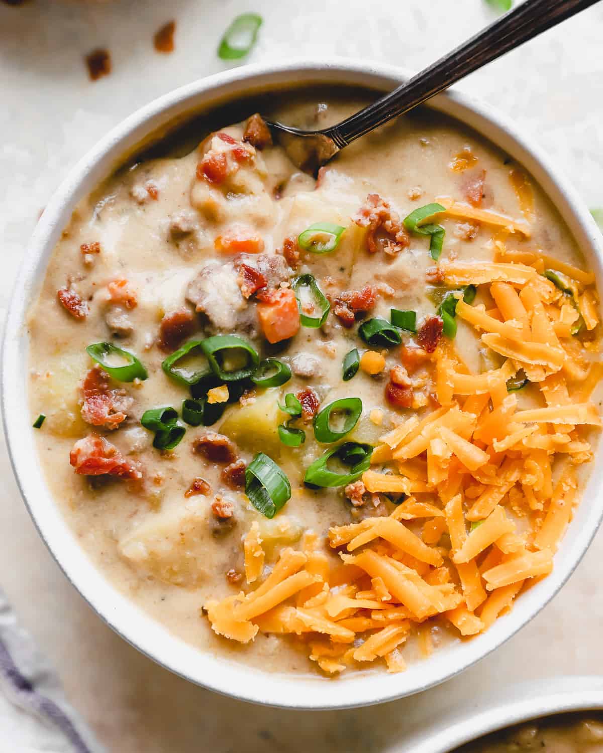 overhead view of a serving of cheeseburger soup in a white bowl with a spoon, topped with bacon, green onion, and shredded cheese.