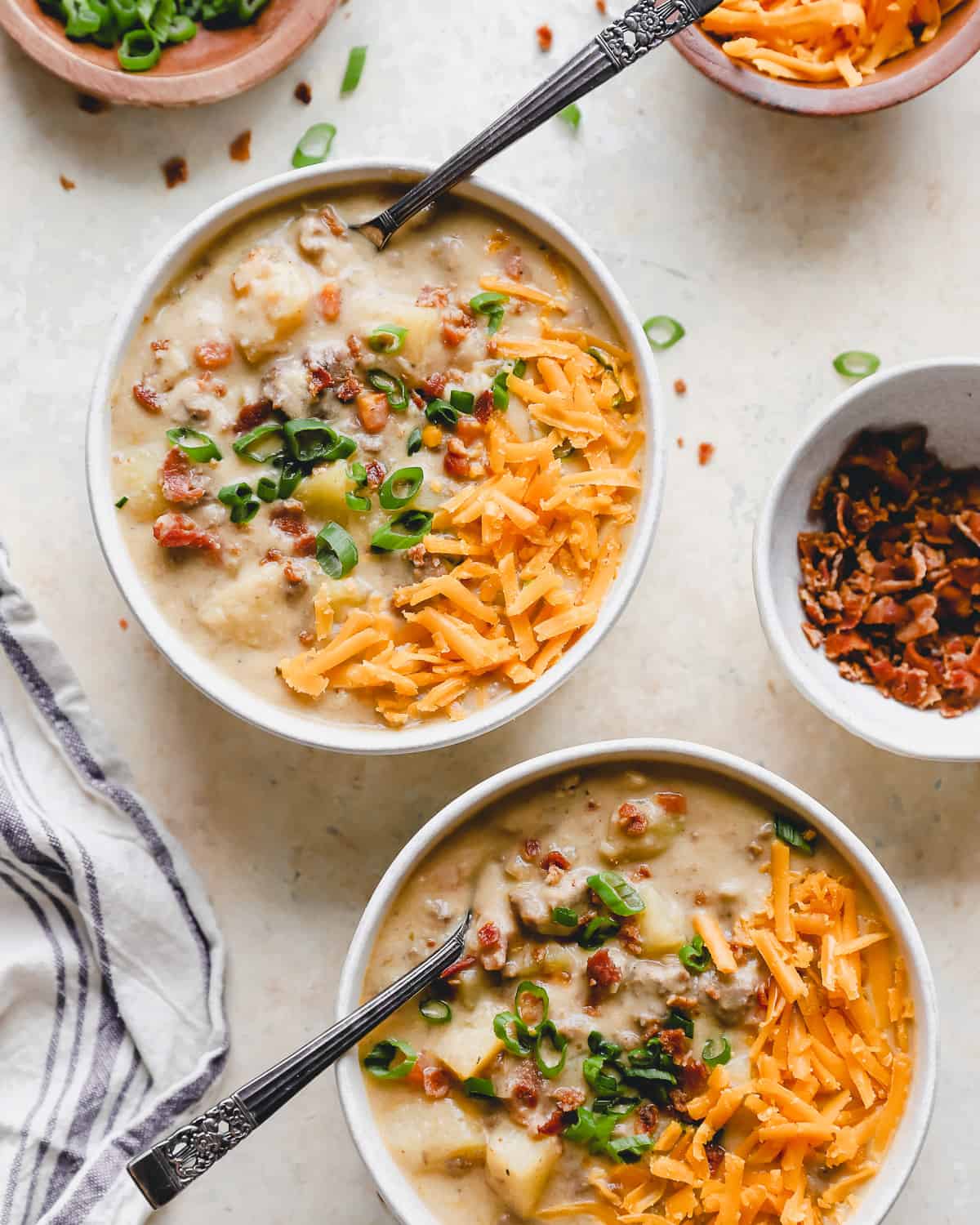 overhead view of 2 servings of bacon cheeseburger soup in white bowls with spoons topped with bacon, green onion, and shredded cheese.