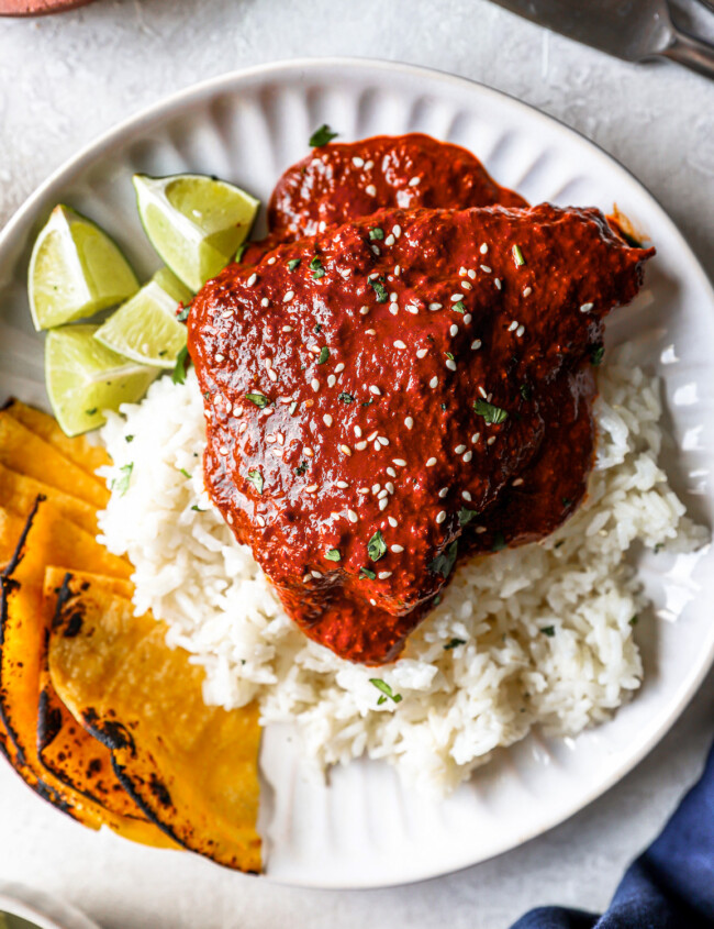overhead view of a serving of chicken mole on a white plate with rice, tortillas, and lime wedges.