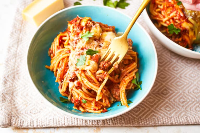 A bowl of spaghetti topped with tomato sauce, grated cheese, and garnished with parsley. A fork is twirling the spaghetti. The bowl is placed on a textured beige cloth, with another bowl of spaghetti and a piece of cheese in the background.