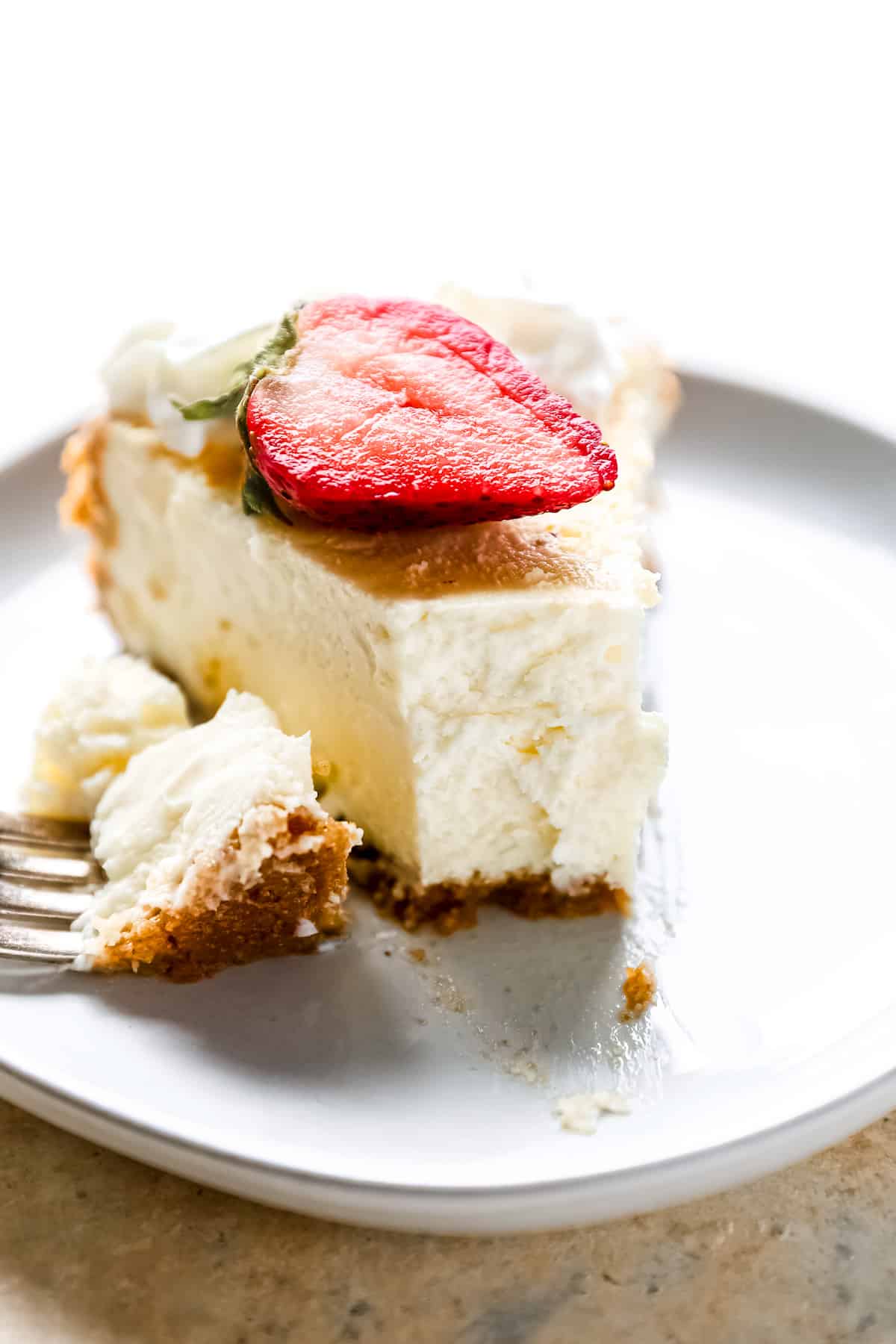 head-on view of a partially eaten slice of easy cheesecake topped with a strawberry on a white plate with a fork.