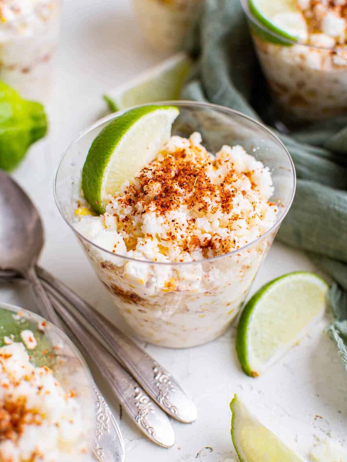 esquites corn in a cup with chili and lime, nestled on a table next to a stack of spoons