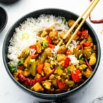 chinese chicken stir fry in a black bowl with rice and chopsticks.