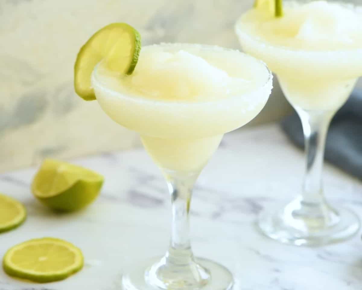 Two frozen margaritas in salt-rimmed glasses garnished with lime wedges and served icy cold.