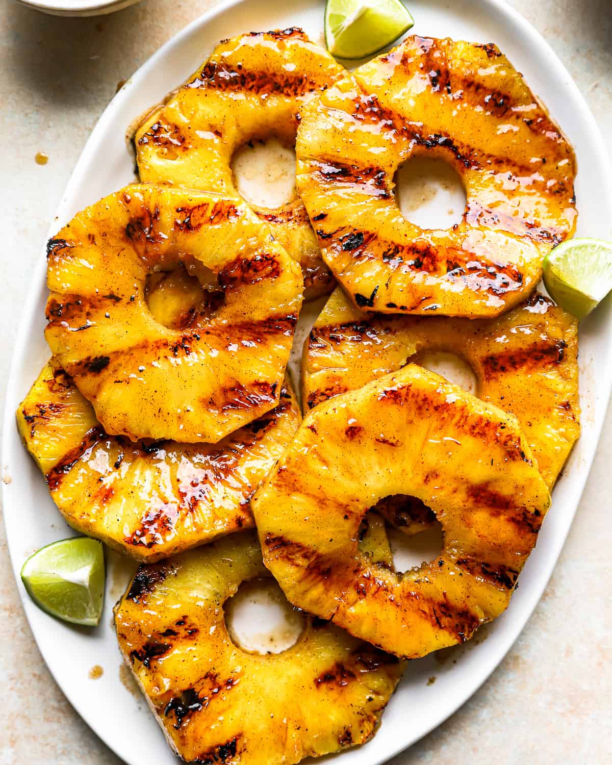 overhead view of slices of grilled pineapple on a white oval serving platter with lime wedges.