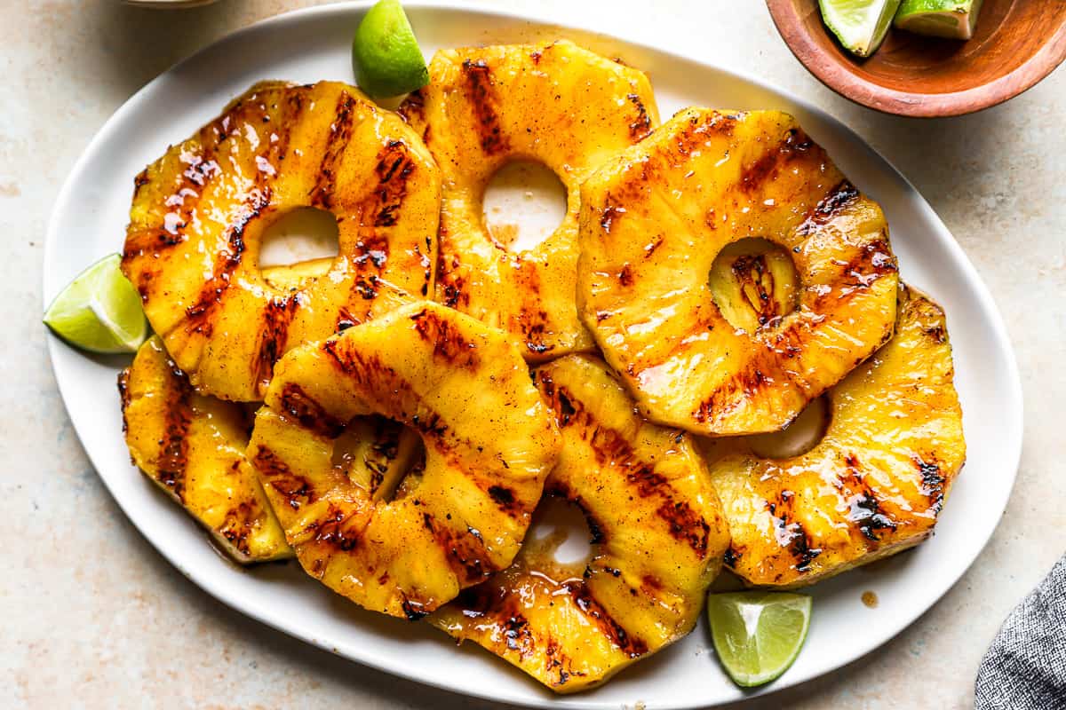 overhead view of a horizontally-positioned oval white platter piled with grilled pineapple slices and lime wedges.