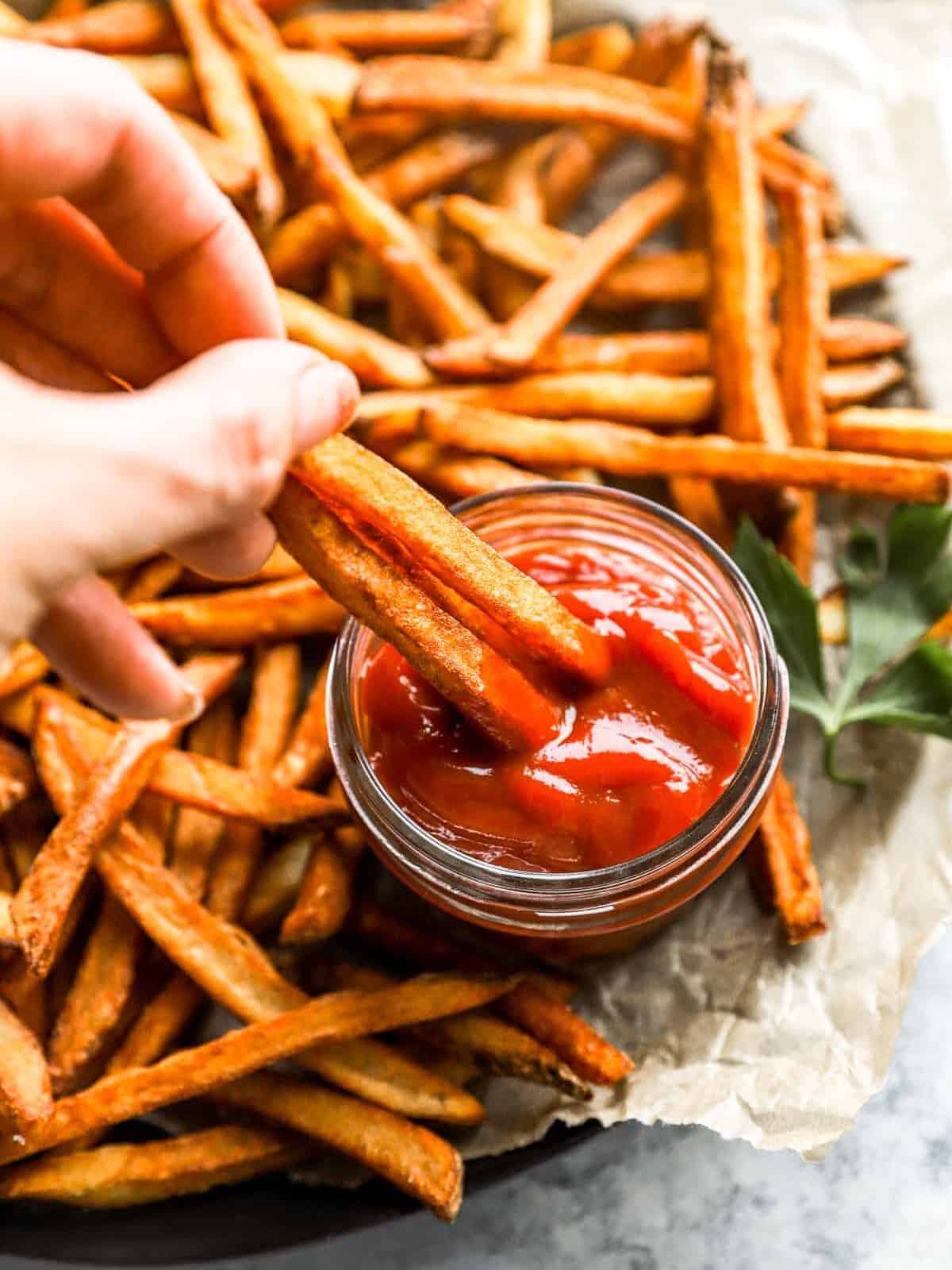 a hand dipping 2 homemade french fries into a cup of ketchup.