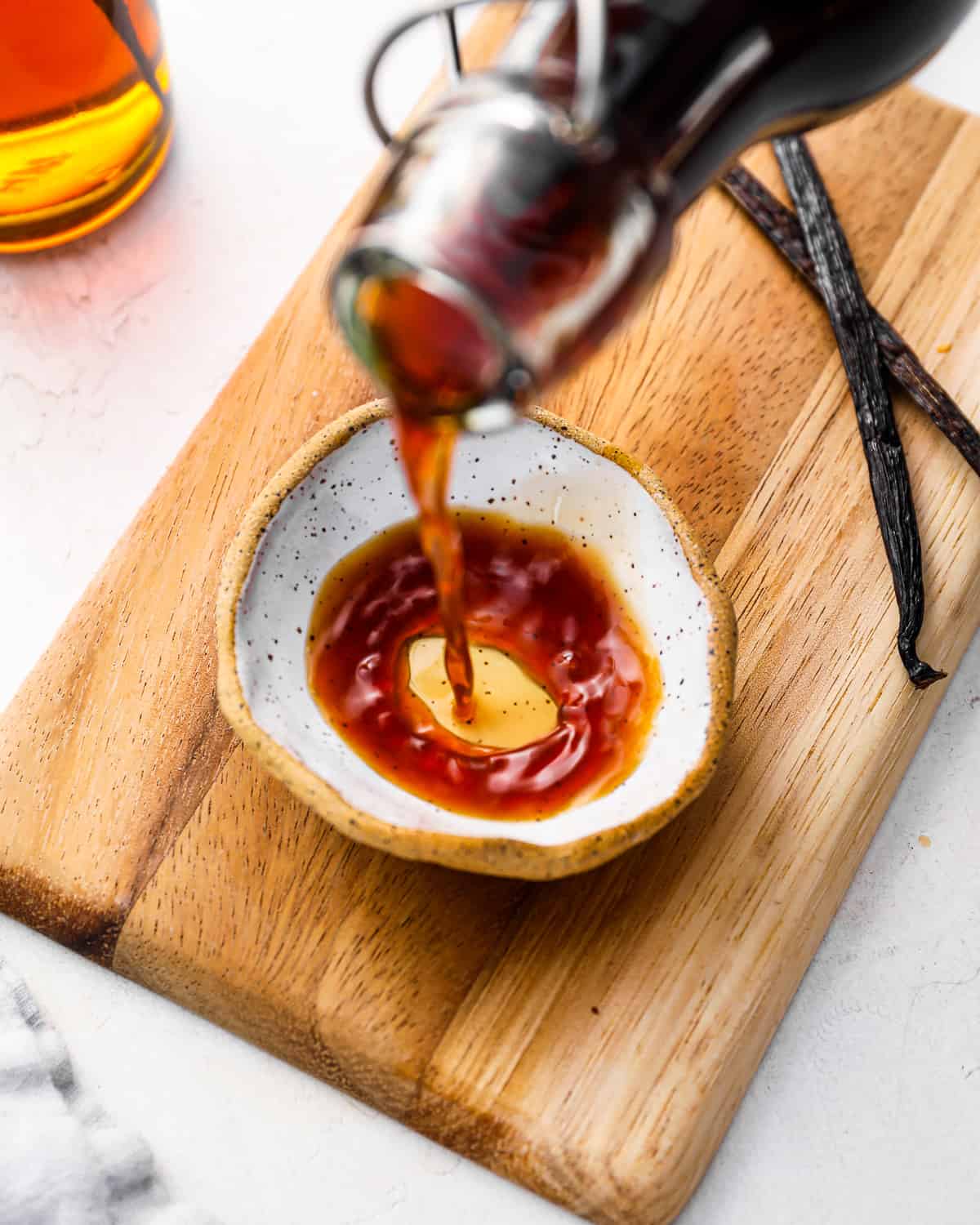 homemade vanilla extract pouring into a small, white dish on a wooden cutting board.