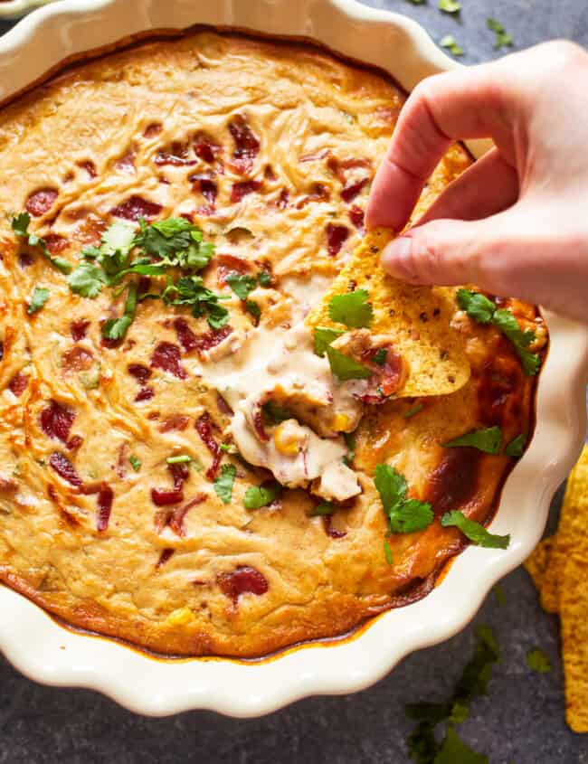 overhead view of a hand dipping a tortilla chip into hot corn and bacon cheese dip in a white round dish.