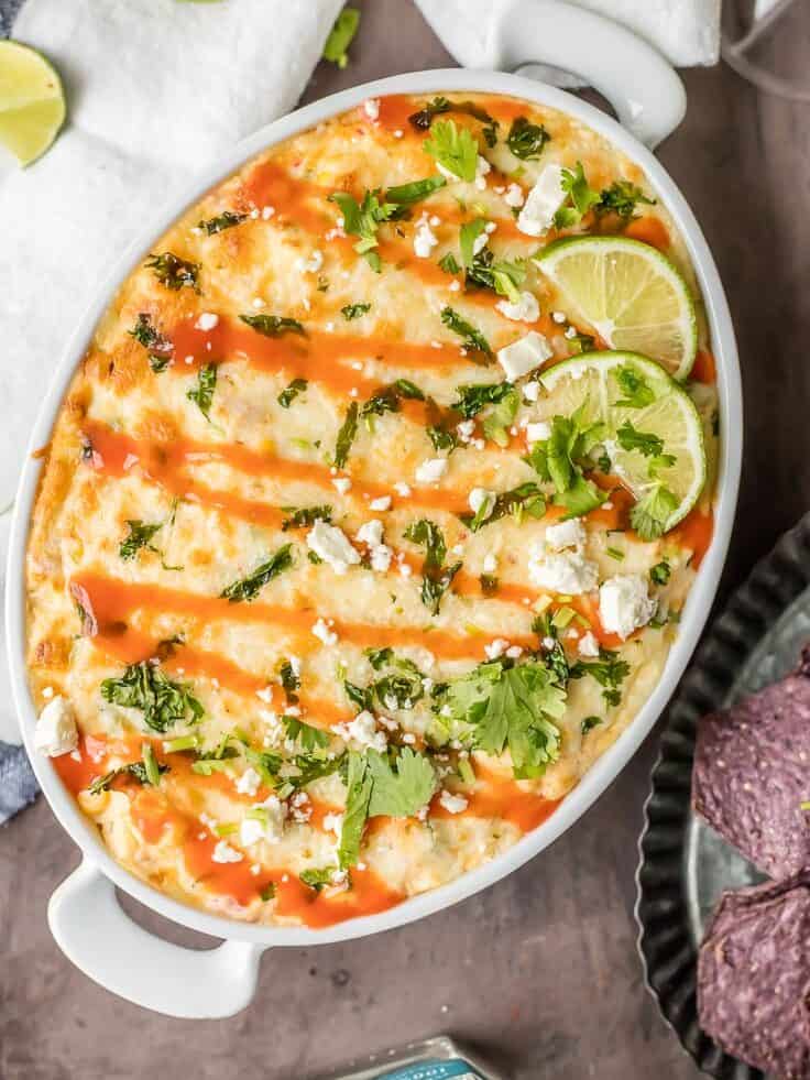 overhead view of spicy mexican corn dip in an oval baking dish, topped with hot sauce, feta, cilantro, and lime 