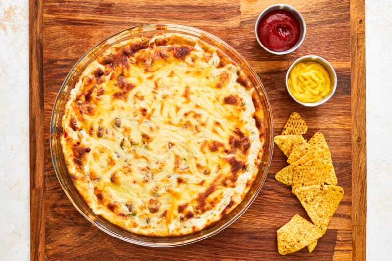 cheeseburger dip in a round baking dish on a wooden cutting board with chips, ketchup, and mustard.