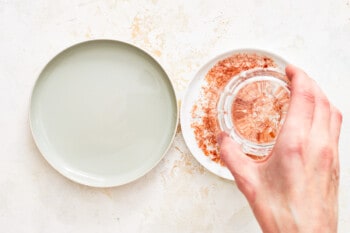 a hand dipping the rim of a glass into chili salt next to a rimmed plate filled with water.