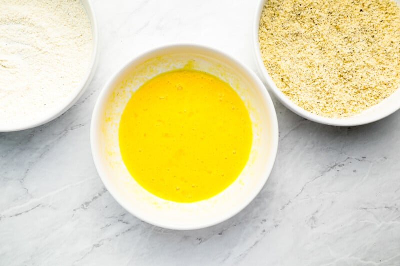 three bowls with flour, eggs and oil on a marble countertop.