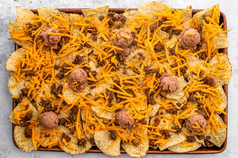 cheese sprinkled over unbaked nachos supreme on a baking sheet.