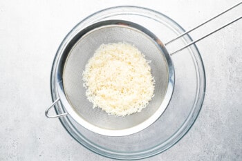 dry basmati rice being rinsed in a fine-mesh sieve set over a glass bowl.