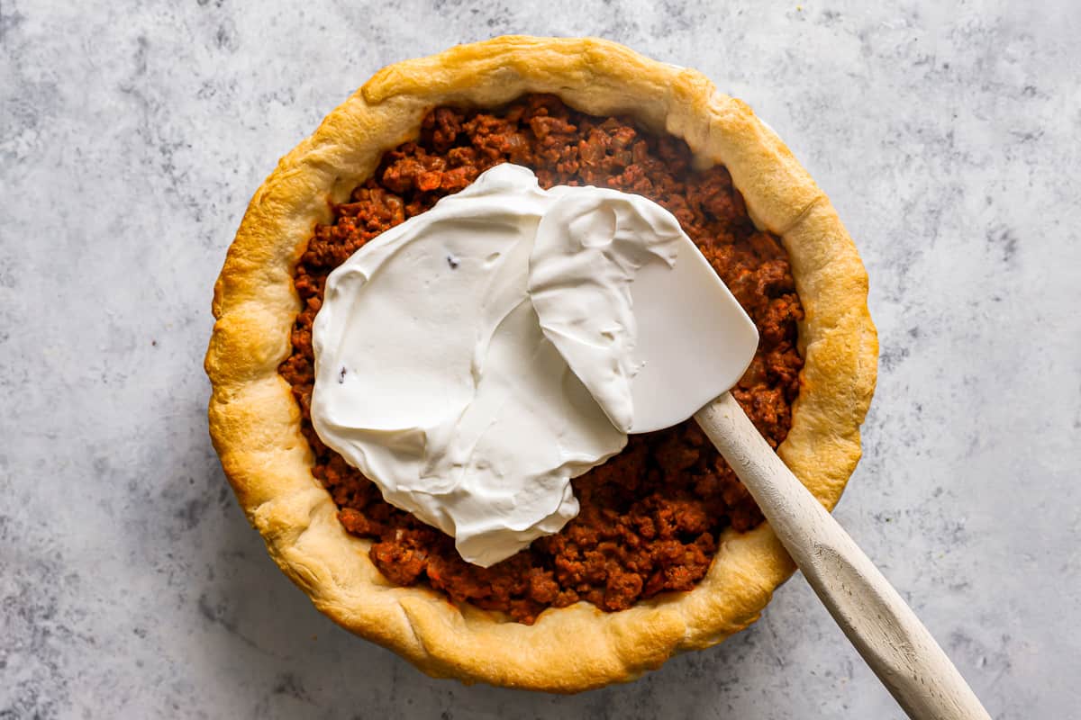 Sour cream being spread over the taco pie in a pie pan with a rubber spatula.