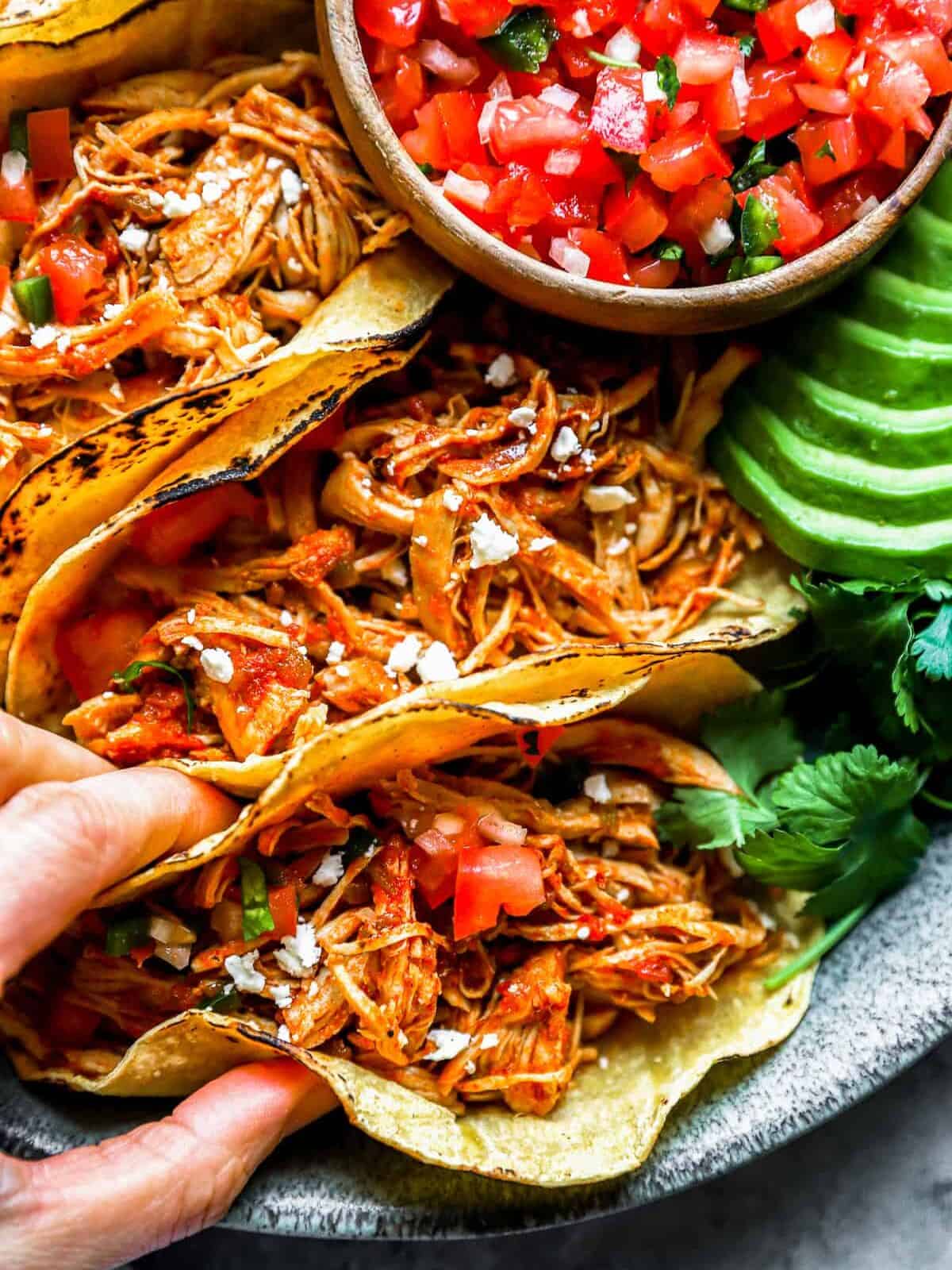 close up overhead view of a hand grabbing 1 of 4 instant pot chicken tacos from a gray plate.